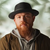 A man with a ginger beard wearing a black hat, grey jumper and green jacket